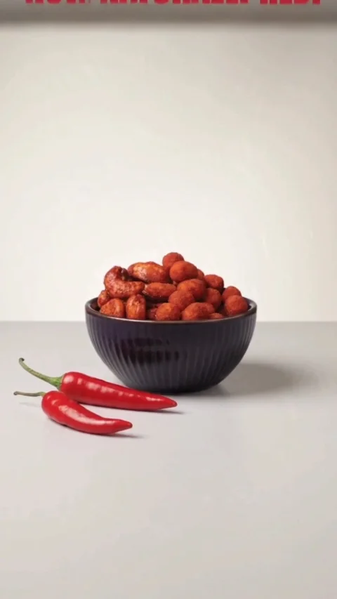 A dark bowl of red-coated peanuts with two red chillies on a pale surface
