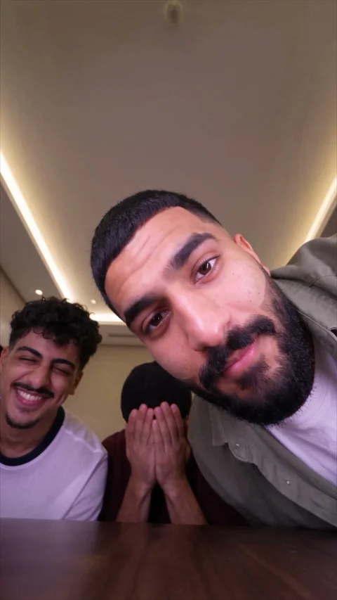 A bearded man leans close to the lens while one friend laughs and another hides his face in his hands, under a lit ceiling