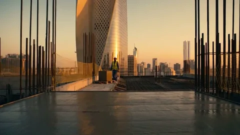 Al Hamra Tower over Kuwait City at sunset, seen from an unfinished concrete rooftop where a worker in a hard hat stands
