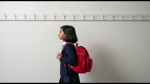 A girl with a red backpack standing in profile against a white wall lined with coat hooks