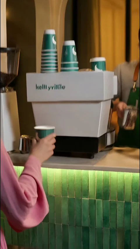 A hand holding a green Kellyville cup at an espresso machine beside a stack of cups