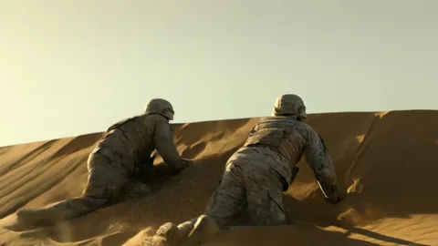 Two soldiers in desert camouflage crawling over the crest of a sand dune