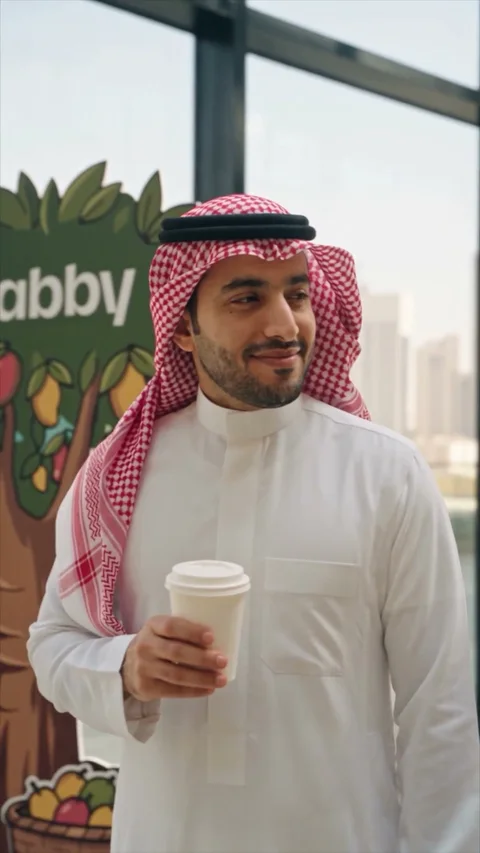 رجل بثوب أبيض وشماغ أحمر يمسك كوب قهوة سفري بجانب لوح على شكل شجرة، وخلفه أبراج المدينة وراء واجهة زجاجية